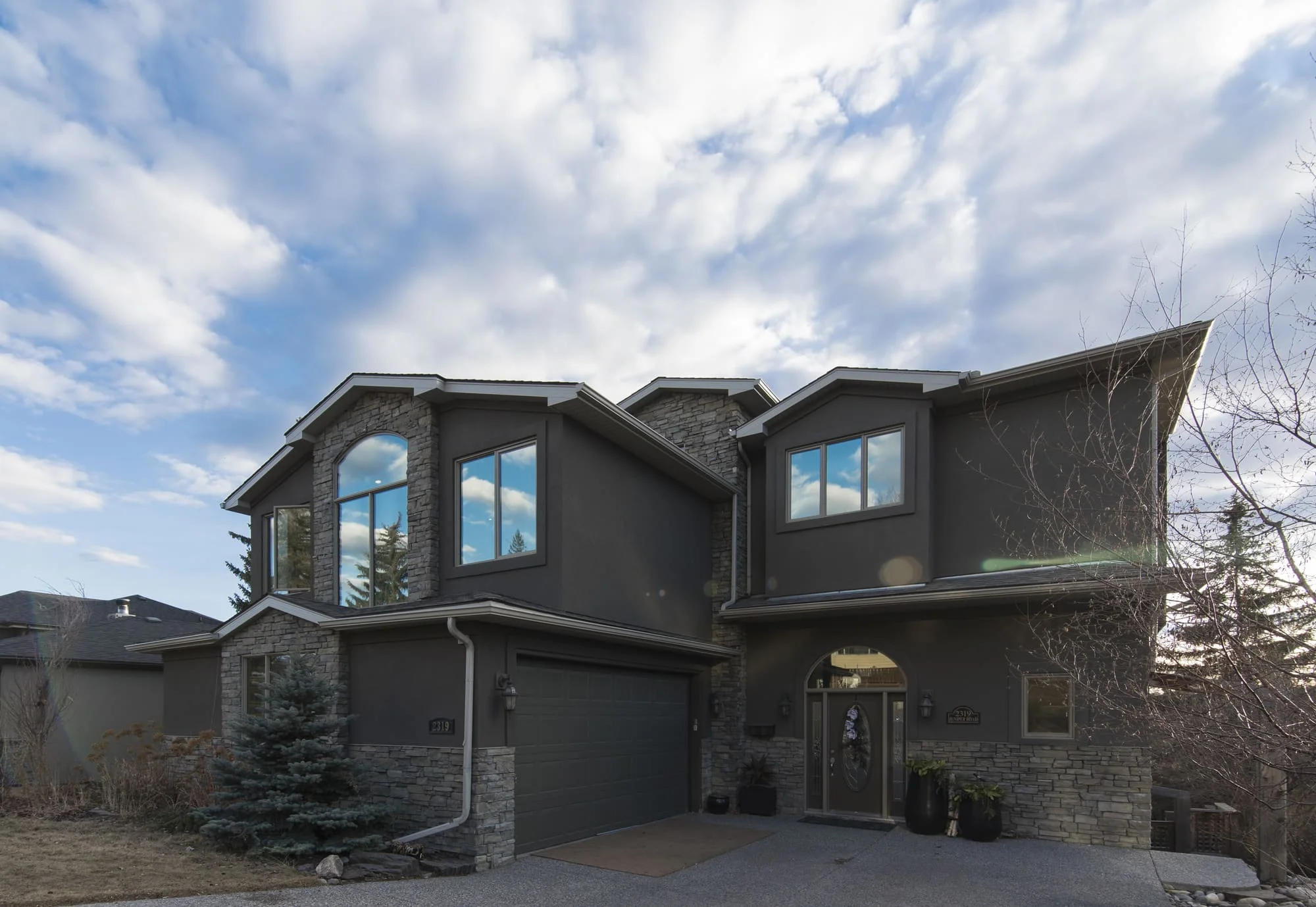 Two-story gray house with a stone base, large windows and a two-car garage under a partly cloudy sky